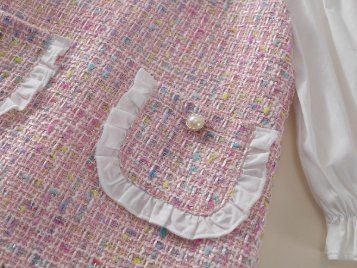 A close view of a tweed pocket with a white ruffle and a pearl button.